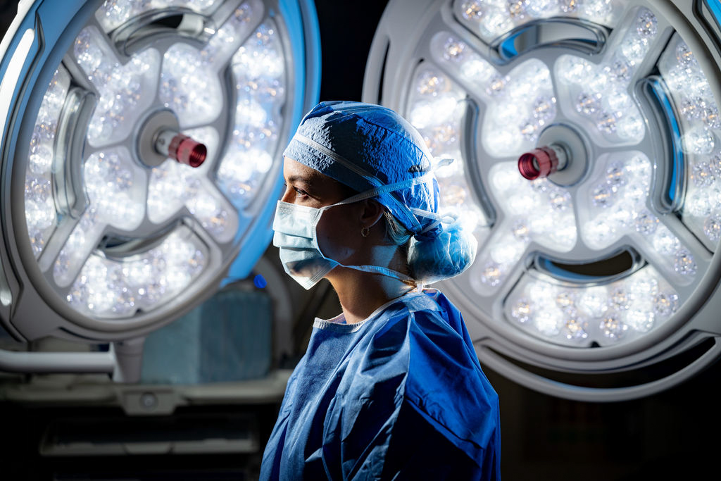 Surgeon wearing surgical scrubs and mask standing in an operating room with bright surgical lights in the background.